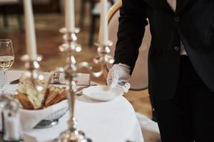 Holds white plate. Process of preparing place for special visitors. Waiter in classical wear works on the servering photo