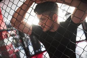 Get ready for exercises. Sportive guy in black shirt stands behind the fence in the room with daylight behind photo