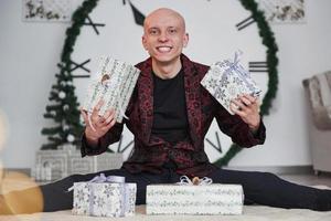 Holding two presents in hands. Man with big clock behind him sitting with New year gift boxes in holiday clothings photo