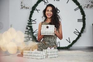 Holding the present. Woman with big clock behind her sitting with New year gift boxes in holiday clothings photo