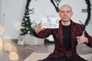 Santa puede estar sin barba y sin pelo. hombre con un gran reloj detrás de él sentado con cajas de regalo de año nuevo en ropa de vacaciones foto