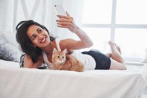 Holding smarthone and taking picture. Attractive blonde resting on the white bed with her cute kitten photo