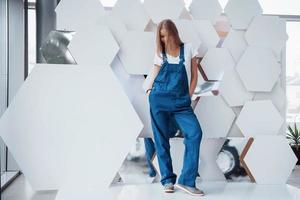 una chica con uniforme azul se alza contra una pared abstracta con piezas blancas de superficie en forma de hexágono en el salón del automóvil foto