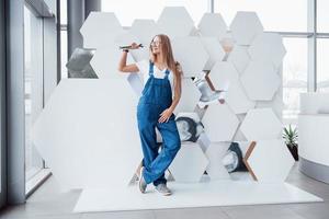 Wrench in hand. Girl in blue uniform stands against abstract wall with white pieces of surface in the shape of hexagon in the auto saloon photo