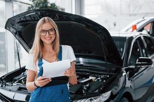 haciendo que sus vehículos rotos vuelvan a ser geniales. gerente con bloc de notas, con camisa blanca y uniforme azul se opone a automóvil con capucha foto