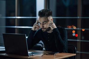 Strong headache. Stylish young businessman works alone in the office at nightime photo