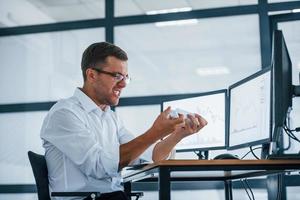 Angry employee throws paper. Conception of failure. Young businessman in formal clothes is in office with multiple screens photo