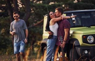 Guy kisses his girlfriend. Group of cheerful friends have nice weekend at sunny day near theirs green car outdoors photo