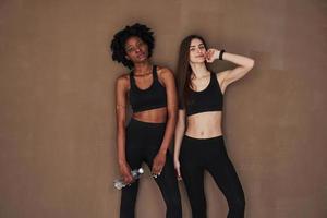 Young and active. Two multi ethnic female friends stands in the studio with brown background photo