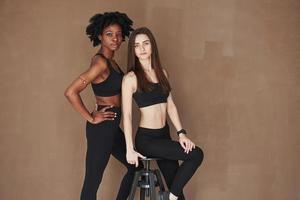 On the black chair. Two multi ethnic female friends stands in the studio with brown background photo