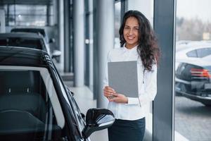 How can I help you. Curly haired female manager stands near the car in automobile salon photo