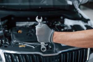 Guantes protectores. la mano del hombre sostiene una llave inglesa frente a un automóvil roto foto