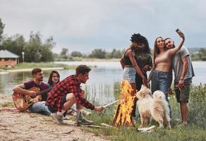 Man puts wood in fire. Group of people have picnic on the beach. Friends have fun at weekend time photo