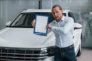 Take a look at this. Manager stands in front of modern white car with paper and documents in hands photo