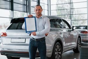 Two vehicles behind. Manager stands in front of modern silver colored car with paper and documents in hands photo