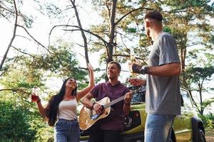 Singing songs together. Friends have nice weekend outdoors near theirs green car with acoustic guitar photo