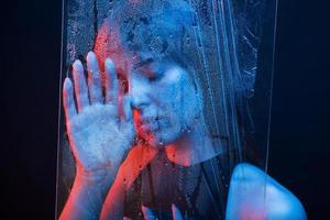 Feel tired. Studio shot in dark studio with neon light. Portrait of beautiful girl behind wet glass photo
