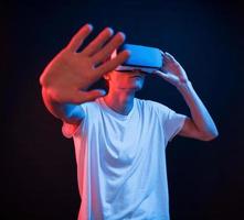 Trying to reach some unreal objects. Young man using virtual reality glasses in the dark room with neon lighting photo