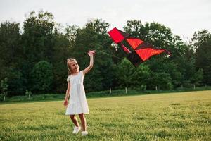 Red and black colored kite. Happy girl in white clothes have fun in the field. Beautiful nature photo