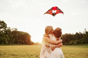 Kid holding kite when mother kissing her. Having fun in the field. Beautiful nature