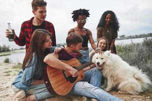 Man plays guitar. Group of people have picnic on the beach. Friends have fun at weekend time photo