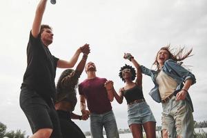 Holding by hands while jumping. Group of people have picnic on the beach. Friends have fun at weekend time photo