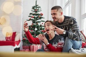 Girl makes happy face for the selfie. Happy young people sits on the windowsill in the room with christmas decorations photo