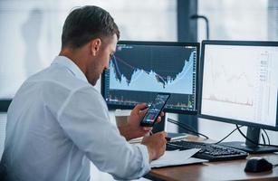 Young businessman in formal clothes is in office with multiple screens. Conception of exchange and money photo