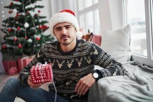 Serious look. Photo of man in santa hat and holiday clothes holds new year gift. Christmas tree on background