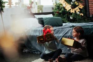 We can't open in right now. Christmas holidays with gifts for these two kids that sitting indoors in the nice room near the bed photo