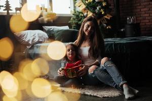 Nice portrait. Mother and daughter sits in holiday decorated room and holds gift box photo