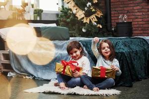 mostrando algún gesto extraño. vacaciones de Navidad con regalos para estos dos niños que están sentados en el interior de la bonita habitación cerca de la cama foto