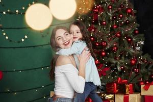 Perfect lighting. Cheerful mother and daughter hugging each other near the Christmas tree that behind. Cute portrait photo