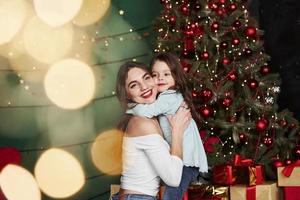 Many of gift boxes on the floor. Cheerful mother and daughter hugging each other near the Christmas tree that behind. Cute portrait photo