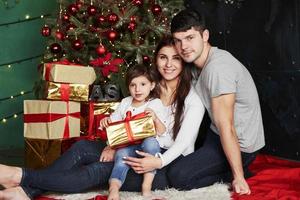 Lovely family sits near the Christmas tree with gift boxes on winter evening, enjoying the time spending together photo