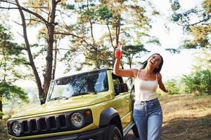 rubia con alcohol en la mano da un paseo por el bosque contra el jeep verde foto