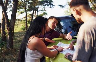 Young friends reading map that is on the hood of modern green jeep in the forest photo