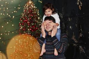 Girl sits on the father's shoulders and covering his eyes. Decorated Christmas at background photo