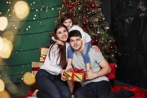 posando para la foto. la encantadora familia se sienta cerca del árbol de navidad con cajas de regalo en la noche de invierno, disfrutando el tiempo que pasan juntos foto