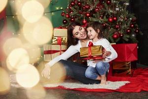 Looking on the side. Cheerful mother and daughter sitting near the Christmas tree that behind. Cute portrait photo