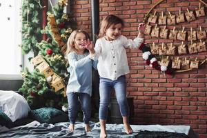 Letters on the tree means merry Christmas. Cheerful kids having fun and jumping on the bed with decorative holiday background photo
