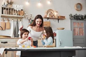 Eating fresh fruits. Young beautiful woman feeds two kids with apples while they sitting near the table with toys photo