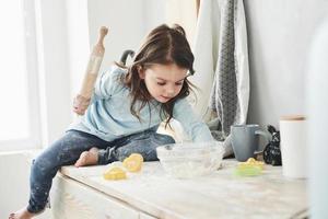 Don't leave your kids alone at home, they can make some food. Photo of pretty little girl that sits on the kitchen table and plays with flour