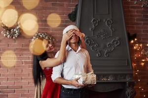 New year gift. Brunette girl close eyes of her man in Santa hat and make him a holiday present photo