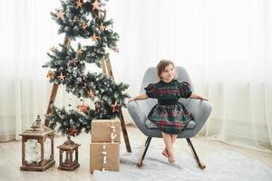 Christmas and holidays conception. Cute little girl is sits on the chair near ladder decorated with stars and gift boxes on the floor photo