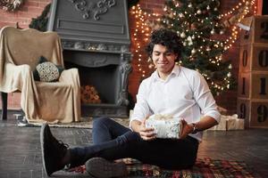 Looking at present with smile. Young man sitting on the floor of beautiful decorated room and holding white gift box in the New year time photo