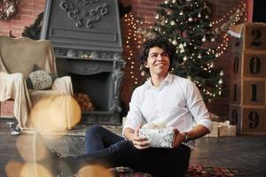 Holiday mood. Young man sitting on the floor of beautiful decorated room and holding white gift box in the New year time photo