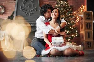 se sienta en el suelo y se abraza. hermosa pareja celebrando el año nuevo en la habitación decorada con árbol de navidad y chimenea detrás foto