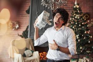 Puedo oír golpear oro dentro. joven parado en la hermosa habitación decorada y sosteniendo una caja de regalo blanca en el año nuevo foto