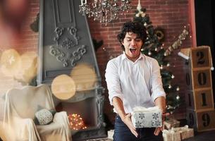 Gifts time. Young man standing in the beautiful decorated room and holding white box with present in the New year time photo